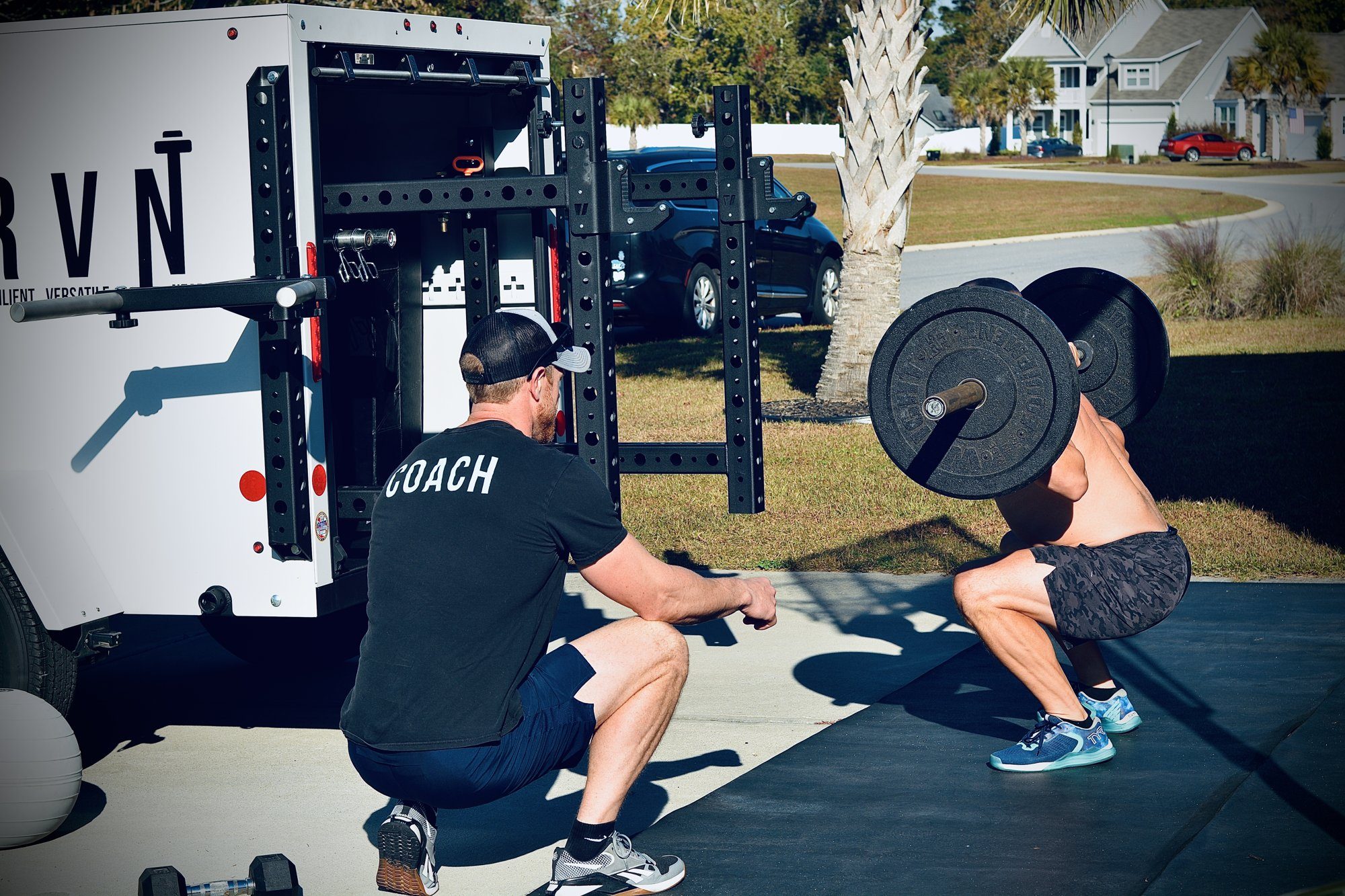 A DRVN coach coaching a client front squat beside the mobile training trailer in a Surfside Beach driveway